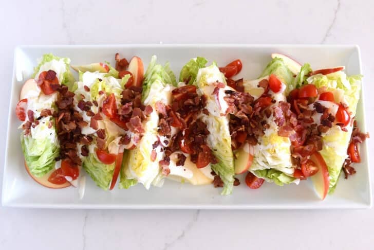 White platter with iceberg wedges, tomatoes, sliced apples, bacon, and blue cheese dressing.