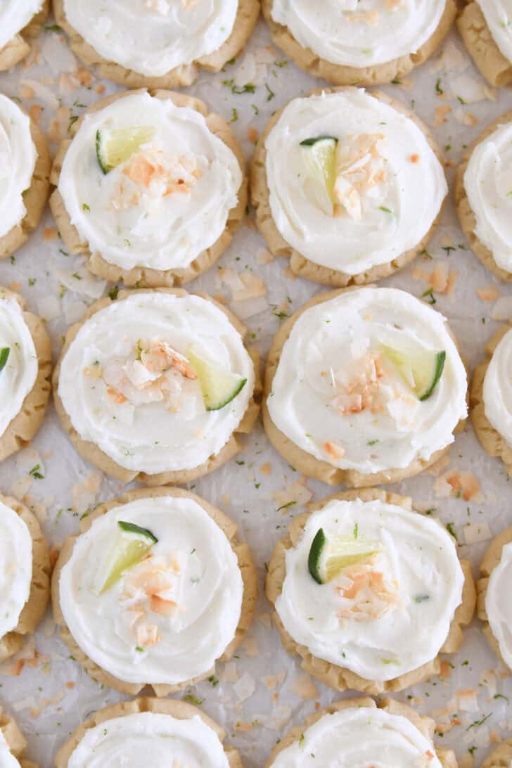 Lots of frosted coconut lime sugar cookies topped with toasted coconut and a fresh lime wedge on white parchment paper.