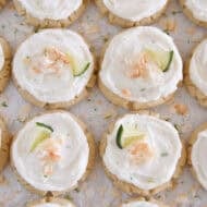 Lots of frosted coconut lime sugar cookies topped with toasted coconut and a fresh lime wedge on white parchment paper.