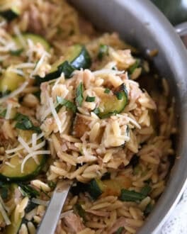 Serving spoon scooping serving of cooked orzo, tuna, zucchini and Parmesan in stainless skillet.