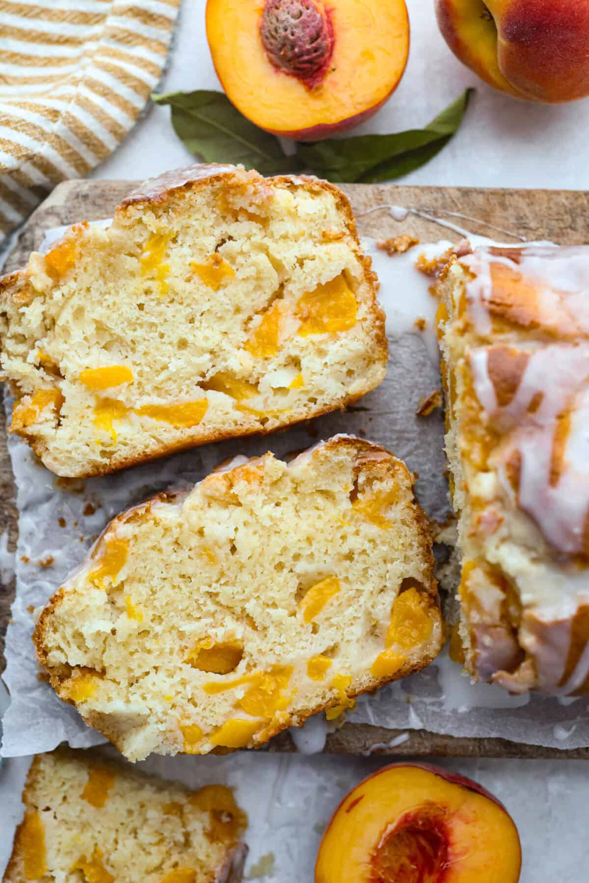 Overhead shot of two slices of peach loaf on a cutting board surrounded by fresh peaches. 