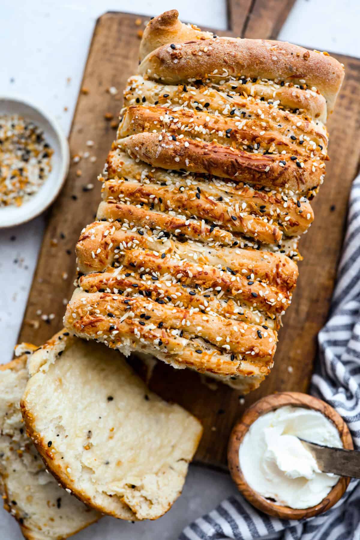 Loaf of everything bagel pull apart bread on a cutting board, sliced with a small bowl of cream cheese next to it. 