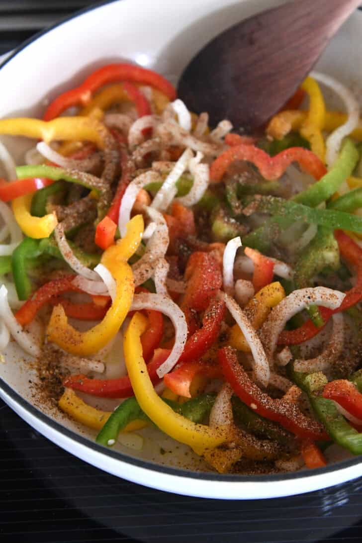 White cast iron skillet with sliced onions, bell peppers and fajita seasonings.