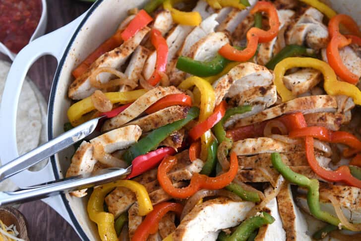White cast iron skillet filled with sliced fajita chicken breasts, bell peppers and onions.