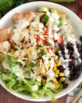 White bowl with cooked chicken, chopped tomatoes, cilantro lime rice, chopped avocados, black beans, roasted corn, and lettuce.