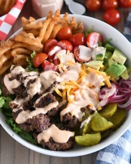 Cooked ground beef, pickles, lettuce, pickled red onions, tomatoes, fries and creamy sauce in white bowl.