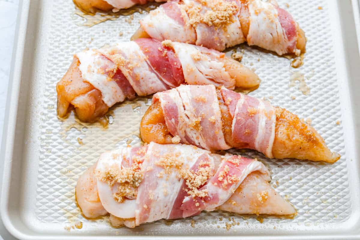 Bacon wrapped chicken tenders on a baking sheet with some more of the brown sugar mixture sprinkled on top. 