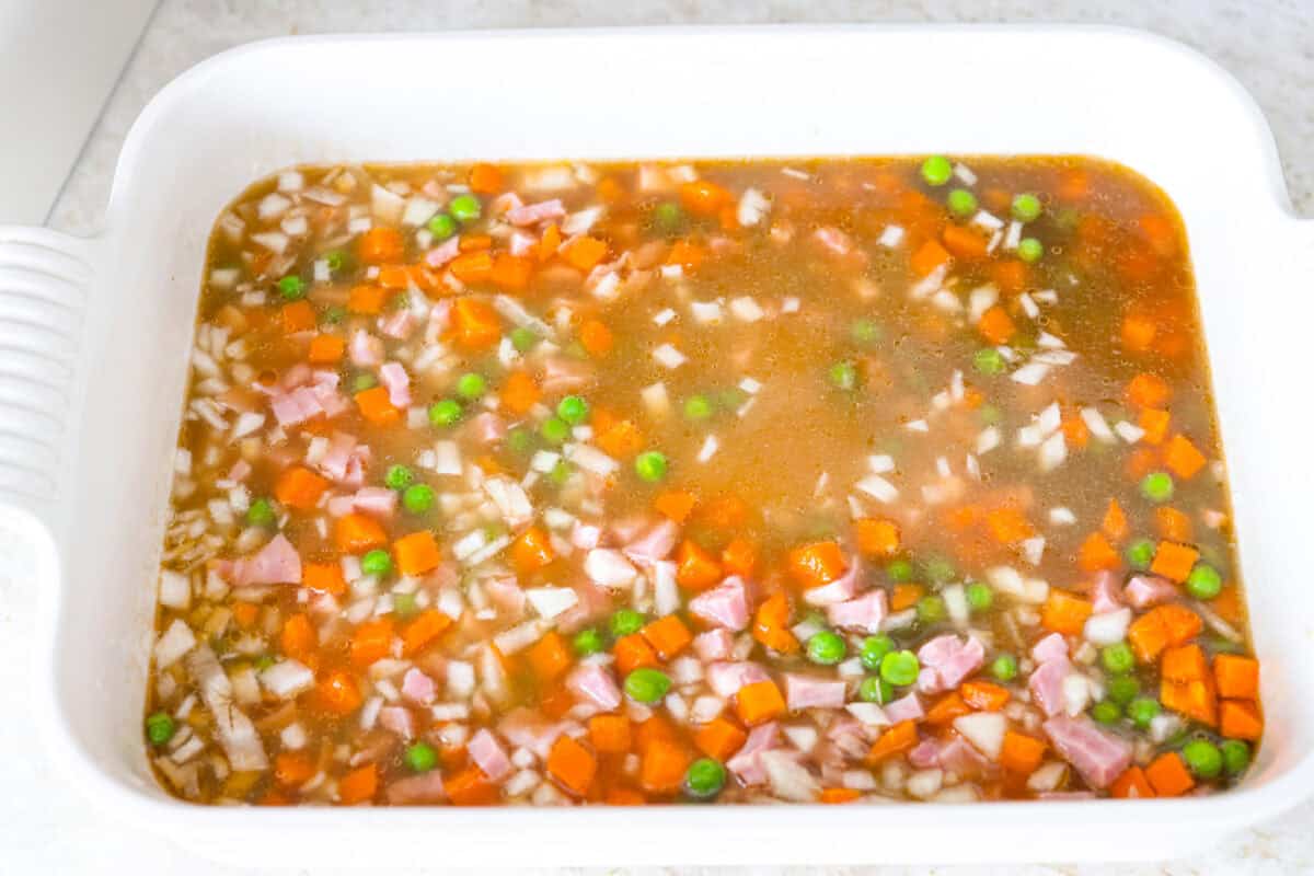 Veggies, ham, rice, garlic, broth and soy sauce added to a baking dish. 