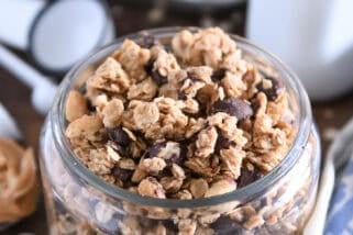 Shallow glass jar filled with peanut butter granola with roasted peanuts and dark chocolate chunks with spoonful of peanut butter to the side.