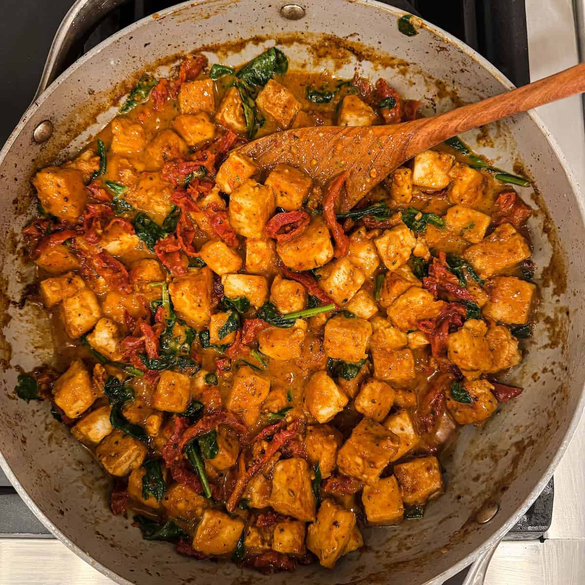Marry me tofu sautéed with sun-dried tomatoes and spinach in a creamy sauce in a skillet with a wooden spoon.